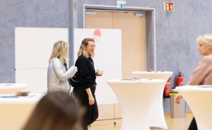 Zwei Frauen stehen in einem Veranstaltungsraum mit weißen Stehtischen und unterhalten sich. Eine hält ein Glas.