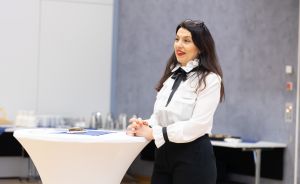 Frau in weißer Bluse mit schwarzer Schleife steht an Stehtisch in Konferenzraum und spricht.