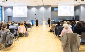 Zwei Frauen präsentieren vor Publikum in einem Konferenzraum, zwei Projektionsleinwände im Hintergrund.