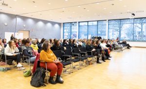 Mehrere Reihen Frauen sitzen auf Stühlen in einem hellen Saal mit großen Fenstern und hören einer Veranstaltung zu.