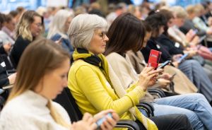 Mehrere Frauen sitzen in Reihen und schauen auf ihre Smartphones. Eine Frau in gelbem Pullover ist im Fokus.
