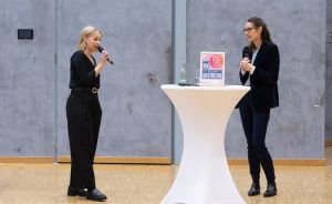 Zwei Frauen mit Mikrofonen stehen an einem weißen Stehtisch in einem Saal und sprechen miteinander.
