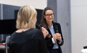 Zwei Frauen mit Mikrofonen im Gespräch; eine mit Brille und dunklem Blazer, eine blonde von hinten sichtbar.