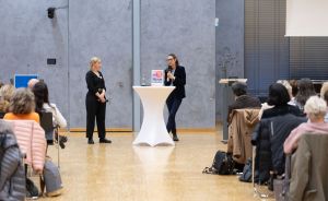 Zwei Frauen stehen vor Publikum in einem Saal. Eine hält ein Mikrofon, zwischen ihnen ein weißer Stehtisch.