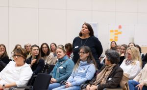 Gruppe von Frauen sitzt in Stuhlreihen; eine Frau steht auf und spricht. Im Hintergrund ein Flipchart mit Notizzetteln.
