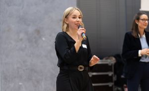 Blonde Frau in schwarzem Jumpsuit spricht in Mikrofon. Im Hintergrund steht eine weitere Frau mit Brille.