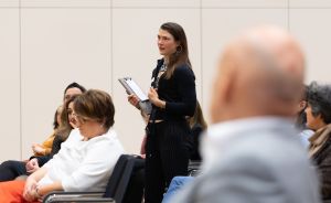 Eine junge Frau steht in einem Konferenzsaal, hält ein Klemmbrett und spricht vor sitzendem Publikum.