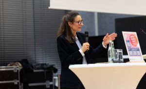 Frau mit Brille und Mikrofon spricht an einem Stehtisch, im Hintergrund ein Plakat und Glasflasche.