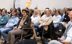 Gruppe von Frauen sitzt in Stuhlreihen in einem Veranstaltungsraum und lacht. Im Hintergrund ein "Welcome Back"-Banner.