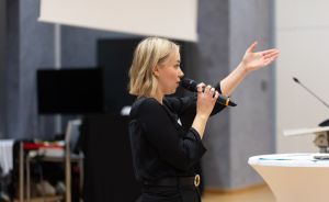 Blonde Frau in Schwarz hält Mikrofon und gestikuliert mit erhobener Hand neben einem Stehtisch.