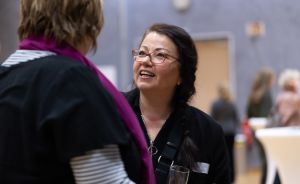 Zwei Frauen im Gespräch bei einer Veranstaltung. Eine lächelt, trägt Brille und hält ein Glas. Hintergrund unscharf.