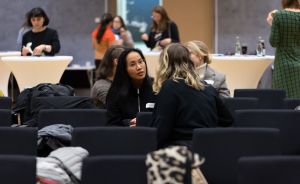 Frauen sitzen in Stuhlreihen eines Konferenzraums und unterhalten sich. Im Hintergrund stehen weitere Personen an Stehtischen.