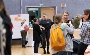 Mehrere Personen stehen in einem Veranstaltungsraum und unterhalten sich. Im Hintergrund ein weißes Pinnwand-Display.
