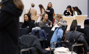 Frauen sitzen auf Stühlen und unterhalten sich in einem Konferenzraum; im Hintergrund stehen weitere Personen an Stehtischen.