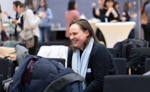 Lächelnde Frau mit hellblauem Schal und dunklem Pullover sitzt in einem Konferenzsaal; im Hintergrund stehen Personen.