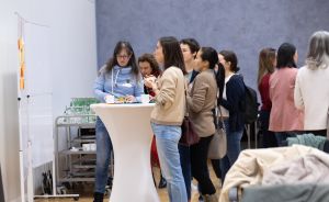 Mehrere Frauen stehen in einem Raum um einen Stehtisch. Im Hintergrund ein Whiteboard und ein Servierwagen.