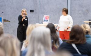 Zwei Frauen stehen vor Publikum. Eine spricht ins Mikrofon, die andere steht daneben. Veranstaltung in einem Saal.