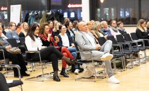 Publikum sitzt auf Stühlen in einer Halle; überwiegend Frauen, ein Mann im grauen Jacket in der vorderen Reihe.