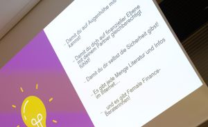 Präsentation auf Leinwand mit deutschen Stichpunkten zu Female Finance, Glühbirnen-Grafik auf lila Hintergrund.