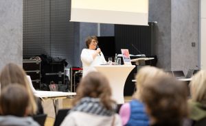 Frau in weißer Kleidung spricht mit Mikrofon an weißem Stehtisch vor sitzenden Zuhörern in einem Saal.