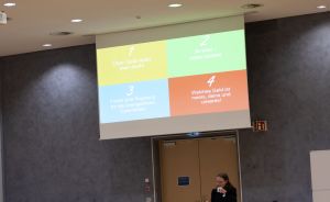 Präsentation auf Leinwand mit 4 farbigen Feldern auf Deutsch, darunter sitzt eine Person an einem Tisch.