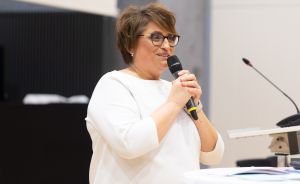 Frau mit kurzen braunen Haaren und Brille spricht in ein Mikrofon, trägt weißes Oberteil.
