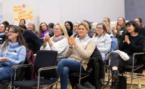 Gruppe von Frauen sitzt in Stühlen und klatscht in einem Veranstaltungsraum. Im Hintergrund ein Whiteboard.