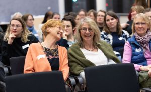Frauen sitzen in Stuhlreihen eines Konferenzraums; zwei Frauen im Vordergrund lachen und tragen Namensschilder.