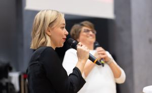 Zwei Frauen halten Mikrofone. Die vordere mit blonden Haaren und schwarzem Oberteil spricht. Die hintere trägt Weiß und Brille.