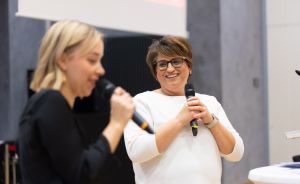 Zwei Frauen stehen nebeneinander und sprechen jeweils in ein Handmikrofon. Eine trägt Schwarz, die andere Weiß.
