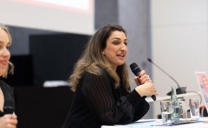 Frau in schwarzer Bluse spricht in Mikrofon an Konferenztisch, neben ihr sitzt eine weitere Person.