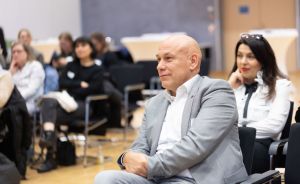 Kahler Mann im grauen Sakko sitzt in einem Veranstaltungssaal, im Hintergrund weitere Personen auf Stühlen.