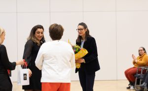 Vier Frauen in einem hellen Raum; eine hält Blumen, eine andere eine Geschenktüte; im Hintergrund klatscht eine Frau.