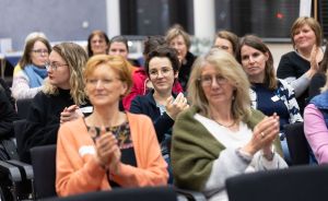 Mehrere Frauen sitzen in Stuhlreihen und klatschen. Sie tragen Namensschilder und schauen nach vorne.