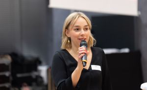 Junge Frau mit blonden Haaren und schwarzer Bluse spricht in ein Mikrofon bei einer Veranstaltung.