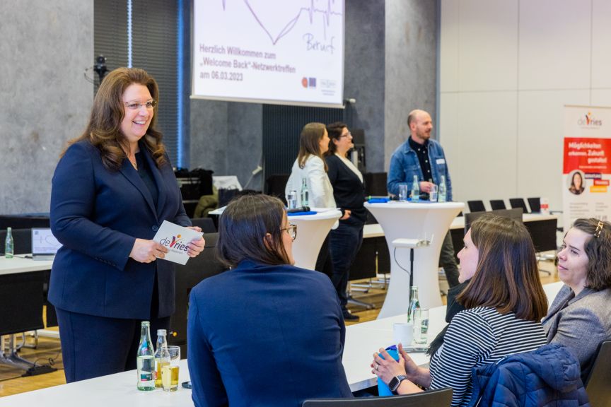 Frau im blauen Blazer spricht mit drei sitzenden Frauen bei einer Netzwerkveranstaltung in einem Konferenzraum.